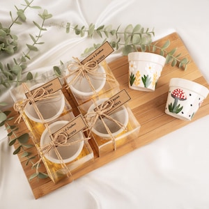 以下が含まれることがあります： 木製のトレイに、花柄やキノコのデザインが施された小さな白い陶器鉢が並んでいます。いくつかの鉢は透明な箱に梱包され、紐で結ばれ、名前と日付が記載されています。ユーカリの小枝が自然な雰囲気を添えています。