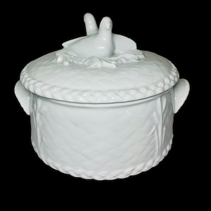 May include: A white ceramic lidded serving dish with a basket weave design. The lid features a pair of doves and a braided edge. The dish has two handles and a textured surface.