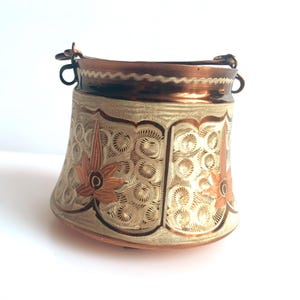 May include: A copper-coloured metal container with a decorative floral and swirl pattern. The container has a handle and a rim. The floral design is in shades of orange and brown. The background is a light beige colour.