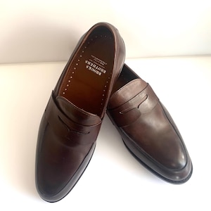 May include: A pair of brown leather penny loafers. The shoes feature a classic design with a rounded toe and a strap across the vamp. The interior of the shoe is brown with the text "BROOKS BROTHERS" printed on the insole.