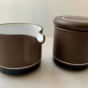 May include: A brown ceramic creamer and sugar bowl set. The creamer has a spout and a white interior. The sugar bowl has a lid. Both pieces have a dark blue band with a white stripe near the base.