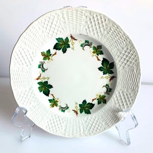 May include: White ceramic plate featuring a textured rim and a central design of green and brown leaves. The plate is displayed on a clear plastic stand, highlighting its decorative pattern. A classic piece for serving or display, suitable for any home.