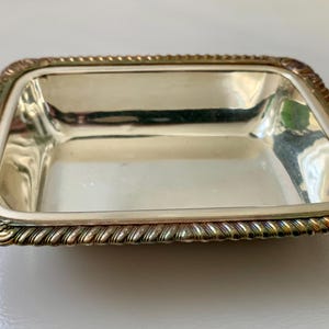 May include: A rectangular silver-plated dish with a decorative rope-like border. The dish has a slightly recessed interior and ornate corner detailing. The silver finish reflects light, creating a bright and elegant appearance. This item is suitable for serving small items.