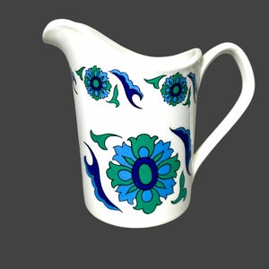 May include: White ceramic jug with a curved spout and handle. The jug is decorated with a floral pattern in shades of blue and green. The flowers are stylized and arranged around the body of the jug.