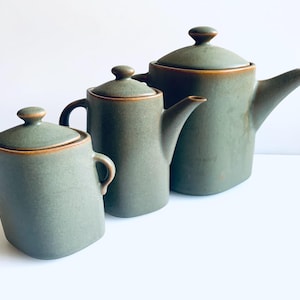 Peut inclure: Un ensemble de trois récipients en céramique vert sauge mat avec des bords bruns. La collection comprend un sucrier avec couvercle et deux anses, une théière moyenne avec couvercle, anse et bec, et une théière plus grande avec couvercle.