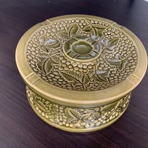 May include: An olive green ceramic ashtray with a lid, featuring a raised grape and leaf pattern. The round ashtray has a small, raised centre and grooves for cigarettes. The design is repeated on the base, creating a decorative piece.