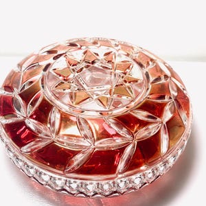 May include: A round, clear glass dish with a decorative, starburst pattern on the lid. The dish has a red and gold colour scheme, with a detailed, cut-glass design. The rim of the dish has a scalloped edge.