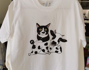 May include: White t-shirt featuring a black and white cat and cow graphic. The cat is sitting on the cow's back. The graphic is a cartoon style. The t-shirt is hanging on a white hanger.