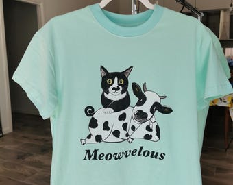 以下が含まれることがあります： 丸首と半袖のライトティールTシャツ。シャツには、黒と白の猫と牛のグラフィックと、黒い文字で「Meowvelous」という言葉が描かれています。シャツは白いハンガーにかかっています。