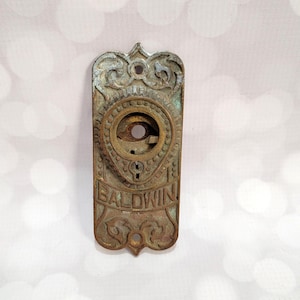 May include: Antique Baldwin door plate with ornate detailing. The rectangular plate is made of aged bronze with a keyhole and the word "BALDWIN" at the bottom. The plate has a decorative top and a circular design around the keyhole.