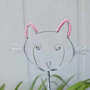 May include: A decorative wire sculpture shaped like a cat head, featuring silver-toned metal wire, pink beads lining the ears, clear beads forming the whiskers, and two small green beads serving as the eyes, all mounted on a single wire stem.