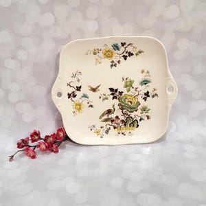 May include: Cream-colored rectangular serving platter with floral and bird design. The platter features yellow, teal, and green flowers with brown branches and a bird. It has handles on each side.