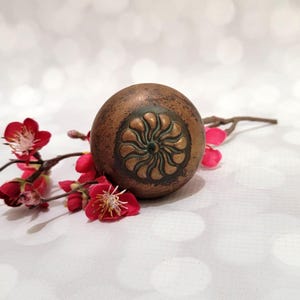 May include: A bronze-colored, spherical decorative object with a floral design on its surface. The object is placed next to a branch with red flowers and buds. The background is a soft, blurred white.
