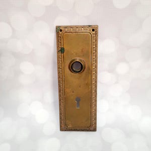 May include: A rectangular antique brass door plate with a keyhole and a circular opening. The plate has an ornate, raised border design. The brass has a vintage patina, showing signs of age and use. The door plate is a vintage architectural element.