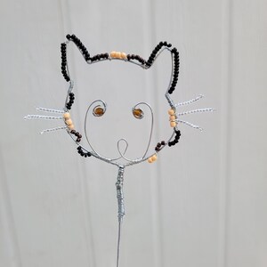 May include: A wire sculpture of a cat head silhouette featuring black and beige beads along the outline, two amber-colored bead eyes, and three silver wire whiskers on each side, all mounted on a single vertical wire stem for display.