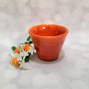 May include: A vibrant orange ceramic planter with a smooth, glossy finish and horizontal ridged detailing along the exterior walls. Beside the planter sits a small decorative cluster of artificial white flowers with orange centers and green leaves.