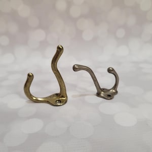 May include: Two metal coat hooks. One is brass-colored, the other is silver-toned. Both have a curved design with a rounded end for hanging items. They are mounted on a surface with a bokeh background.