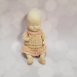May include: A small, vintage doll with a blank face, wearing a crocheted dress and matching shoes. The dress is beige with pink trim around the neckline and hem. The doll's arms and legs are also visible.