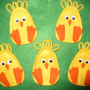 May include: Five yellow felt chick ornaments with orange feet and white eyes. The chicks are sitting on a green felt background.