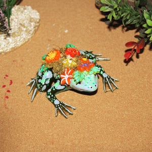 May include: A decorative frog figurine with a light blue body, black spots, and silver-colored feet. The frog is adorned with colorful flowers and greenery on its back. The figurine is set against a brown background with decorative plants.