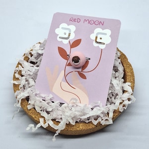 Puede incluir: Un anillo rosa con un centro marrón se muestra en una tarjeta lavanda con el texto "Red Moon". La tarjeta presenta un diseño floral y está colocada en un plato de corcho con papel blanco.