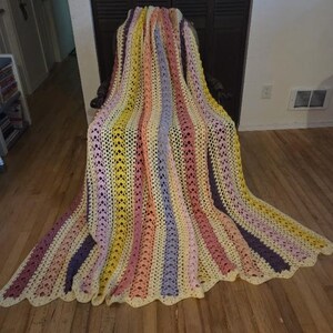 May include: A handmade, colorful crochet blanket draped over a chair. The blanket features vertical stripes in shades of yellow, pink, purple, and cream. The edges have a scalloped design. The blanket is made of soft yarn.