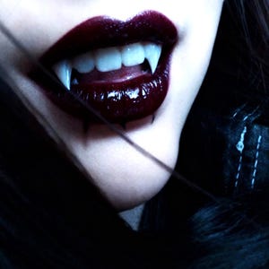 May include: Close-up of a person's mouth with dark burgundy lipstick and sharp, white fangs. The lips are glossy, and the skin is pale. Dark hair frames the face, creating a gothic or vampire-themed aesthetic.