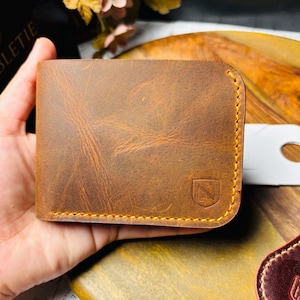 Puede incluir: Cartera de cuero marrón con textura envejecida. La cartera tiene un borde cosido y un pequeño emblema. Se muestra sobre una superficie de madera, sostenida en una mano.