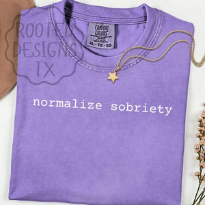 May include: A lavender Comfort Colors t-shirt with the phrase "normalize sobriety" printed in white. A gold star pendant necklace rests on the shirt. The shirt is folded and has a tag that reads XL-TG-EG.