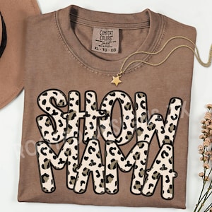 Op de afbeelding: Een bruin Comfort Colors T-shirt met de woorden "SHOW MAMA" in een luipaardprint ontwerp. Een gouden sterrenhangerketting ligt op het shirt. Het shirt is opgevouwen en heeft een label met XL-TG-EG.