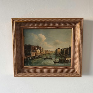 May include: An antique framed painting depicting a canal scene with gondolas and buildings. The artwork features a gold-colored ornate frame. The painting showcases a detailed landscape with a blue sky and clouds, evoking a sense of historical charm.