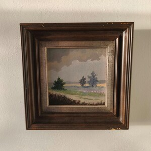 Op de afbeelding: Een ingelijst landschapsschilderij met een veld met bomen onder een bewolkte lucht. Het schilderij heeft een donkerbruine houten lijst. Het kunstwerk toont een serene buitenscène met gedempte kleuren.