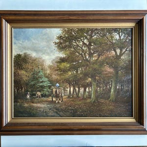 Op de afbeelding: Een ingelijst olieverfschilderij dat een landelijke scène toont met bomen, figuren en een paardenkar. Het kunstwerk toont een gedetailleerd landschap met een bewolkte lucht en herfstkleuren. De lijst is donkerbruin met een gouden binnenrand.