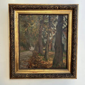 Puede incluir: Una pintura al óleo que representa un camino bordeado de árboles cubierto de hojas de otoño, enmarcada en un marco de madera dorado oscuro ornamentado con intrincadas tallas florales en los bordes. La escena muestra troncos oscuros.