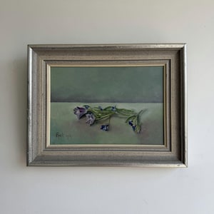 May include: An oil painting in a silver-toned frame depicting a small cluster of purple and blue bell-shaped flowers resting on a flat surface against a muted green background. The artist signature Riet 1977 is visible in the lower left corner.