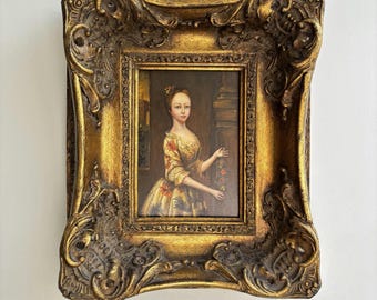 May include: An oil painting portrait of a person wearing a yellow dress with floral patterns, set within a heavy, ornate, gold-colored frame featuring intricate scrollwork and relief details. The subject stands in a dimly lit interior space.