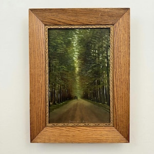 Puede incluir: Un óleo enmarcado que representa un camino de tierra bordeado por altos árboles verdes, encerrado en un marco ancho de madera de roble texturizada con un borde interior fino, dorado y ornamentado. La escena muestra un sendero forestal.