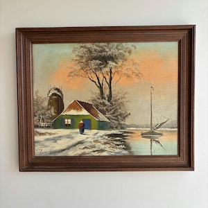 Op de afbeelding: Een ingelijst olieverfschilderij met een winterlandschap met een groen huis, een persoon in een rode jas, een hoge boom en een zeilboot op kalm water. De lucht heeft een zachte oranje en blauwe kleurverloop en de grond is bedekt met witte sneeuw.