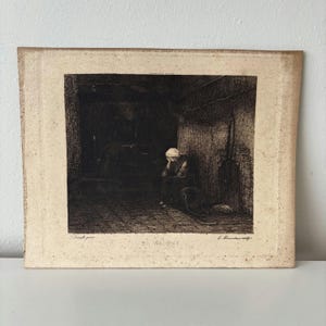 Op de afbeelding: Een vintage prent die een persoon toont die in een zwak verlicht interieur zit. Het kunstwerk, in sepiatinten, toont de tekst "DE WEDUWE" en de handtekening van de kunstenaar. De prent is gemonteerd op verouderd papier.