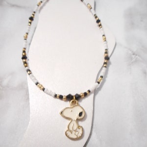 以下が含まれることがあります： 白、黒、金のビーズチェーンのアンクレット。 金色のスヌーピーチャームがチェーンから吊り下げられています。 チャームは白地に黒のディテール。 アンクレットは白い表面に表示されています。