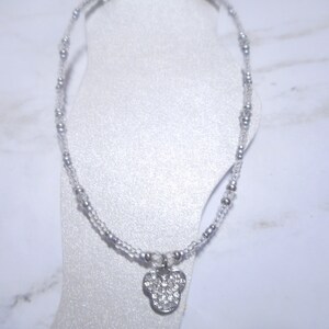 May include: A silver anklet with a Mickey Mouse pendant. The anklet features small, clear beads and larger silver beads. The pendant is shaped like Mickey Mouse's head and is encrusted with clear rhinestones. The anklet is displayed on a white foot form.