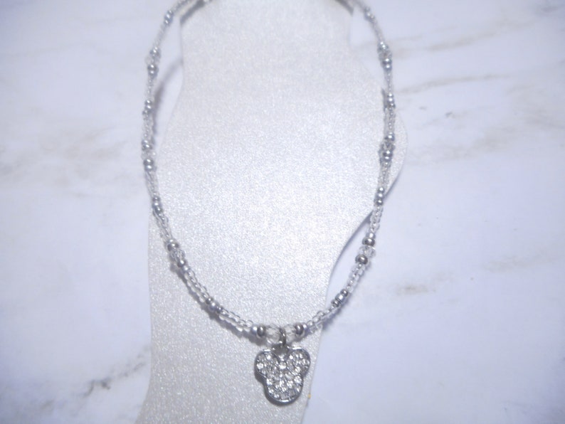 May include: A silver anklet with a Mickey Mouse pendant. The anklet features small, clear beads and larger silver beads. The pendant is shaped like Mickey Mouse's head and is encrusted with clear rhinestones. The anklet is displayed on a white foot form.