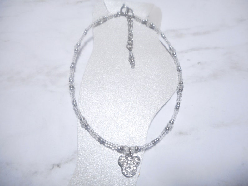 May include: A silver anklet with a clear bead strand and small silver beads. The anklet features a pendant in the shape of a mouse head, encrusted with clear rhinestones. The anklet is displayed on a white foot form.