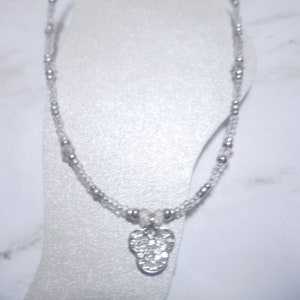 May include: A delicate anklet featuring a clear beaded chain with small silver-colored beads. A silver-colored charm in the shape of a flower is suspended from the chain. The anklet is displayed on a white surface.