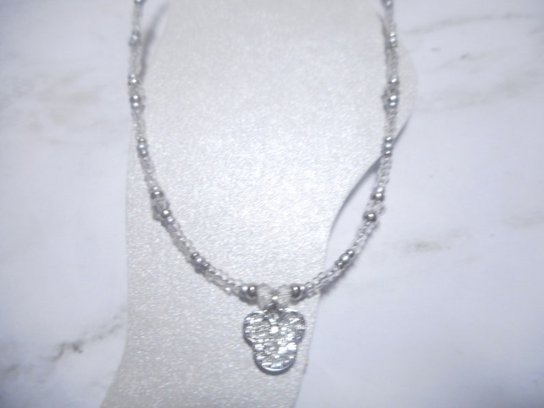 May include: A delicate anklet featuring a clear beaded chain with small silver-colored beads. A silver-colored charm in the shape of a flower is suspended from the chain. The anklet is displayed on a white surface.