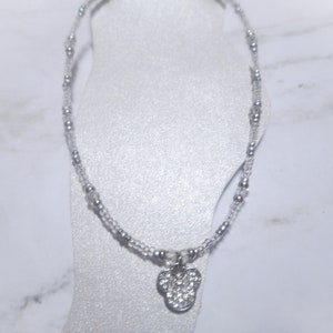 May include: A delicate silver anklet featuring a small, sparkling charm in the shape of a mouse head. The anklet is made of small, clear beads and silver accents, creating a subtle and elegant look.