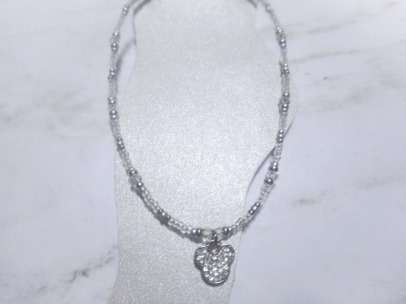 May include: A delicate silver anklet featuring a small, sparkling charm in the shape of a mouse head. The anklet is made of small, clear beads and silver accents, creating a subtle and elegant look.