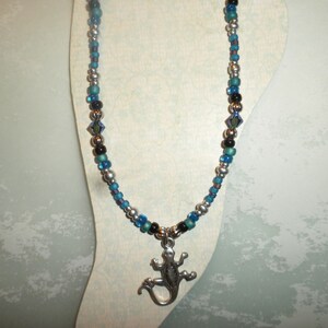 May include: A beaded necklace with a silver lizard pendant. The necklace features a mix of silver, blue, and black beads. The lizard pendant is detailed and adds a unique touch to the jewelry.