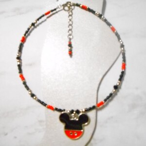 Pode incluir: Tornozeleira com um pingente do Mickey Mouse. A corrente de contas &eacute; preta, vermelha e dourada. O pingente do Mickey Mouse &eacute; preto com um rosto vermelho e branco. A tornozeleira &eacute; exibida em uma superf&iacute;cie branca.