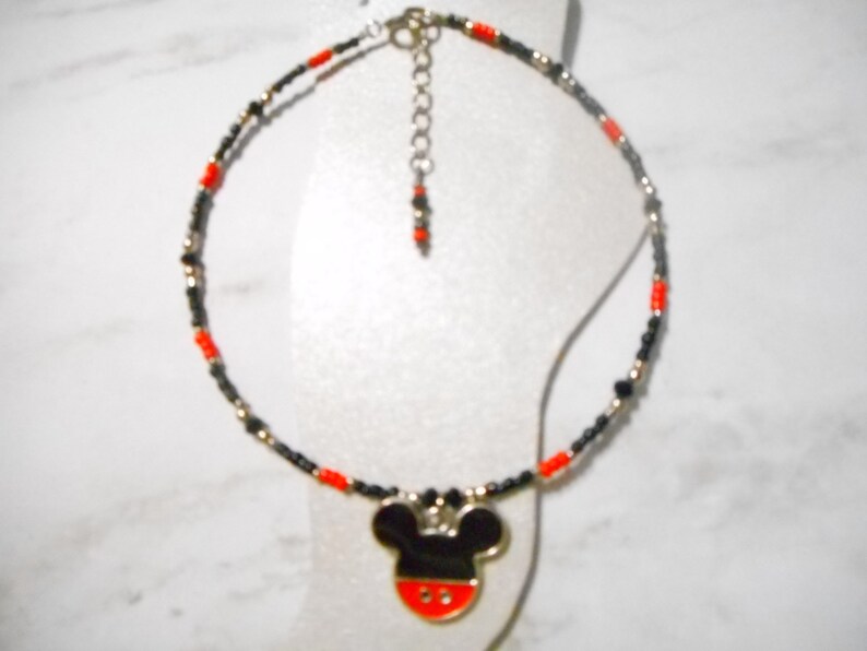 Pode incluir: Tornozeleira com um pingente do Mickey Mouse. A corrente de contas &eacute; preta, vermelha e dourada. O pingente do Mickey Mouse &eacute; preto com um rosto vermelho e branco. A tornozeleira &eacute; exibida em uma superf&iacute;cie branca.